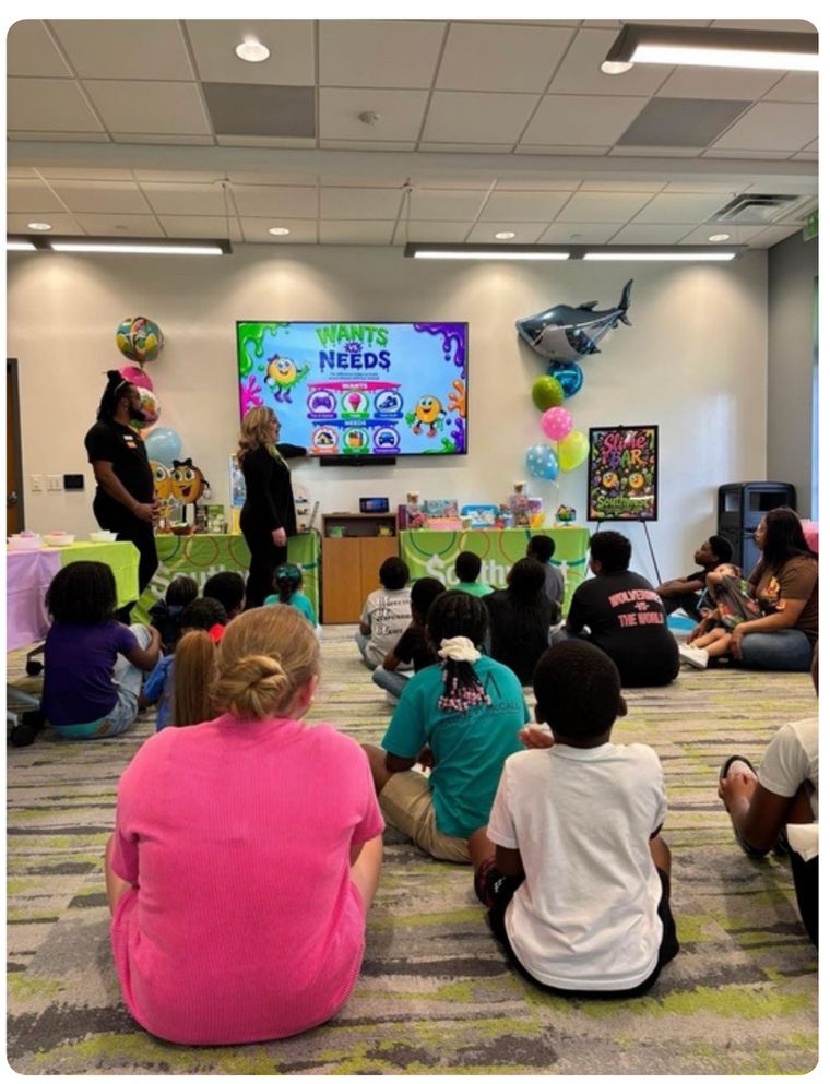 A room full of children seated on the floor watching a Wants vs. Needs presentation on a large screen at a partner branch event