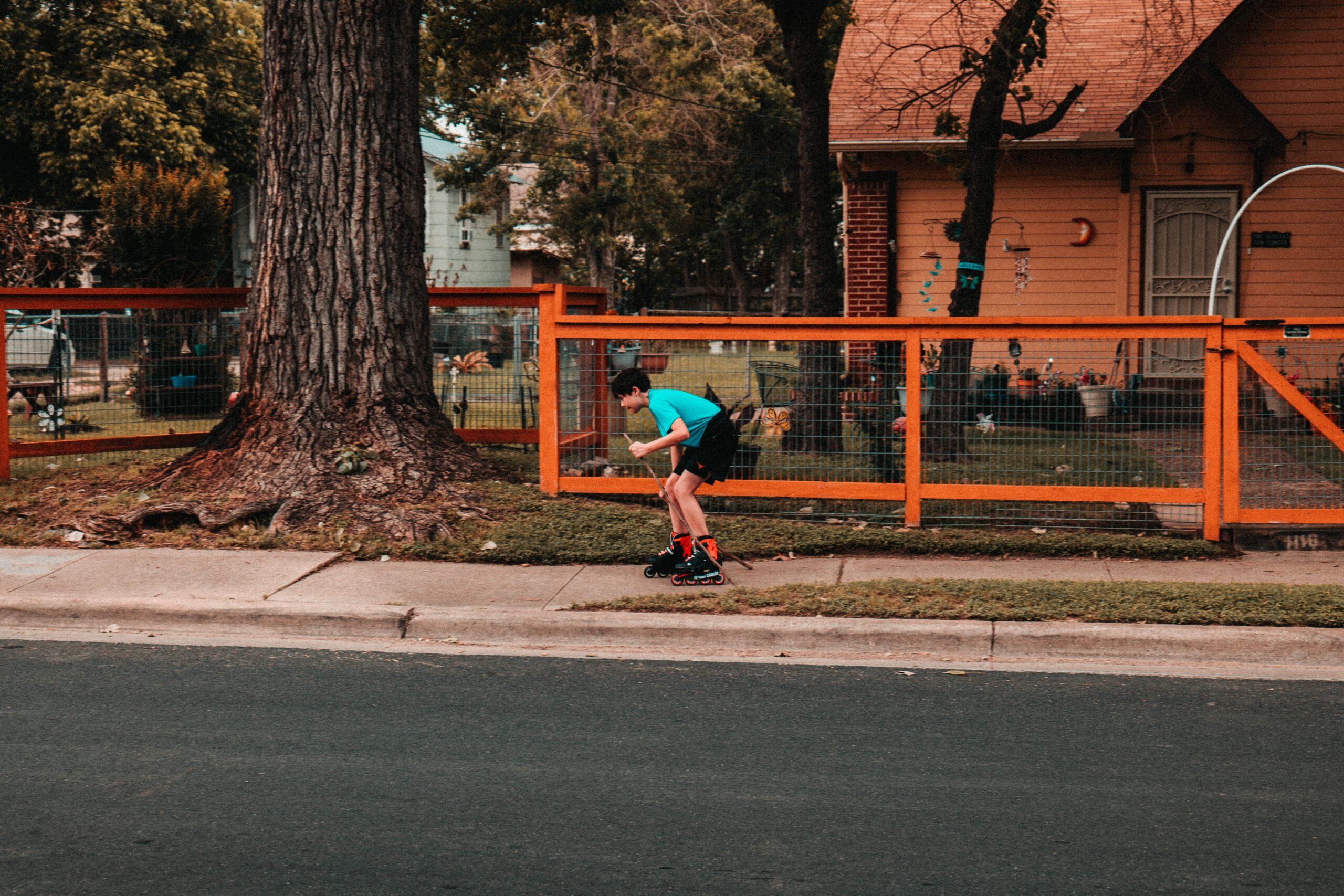 A boy rollerblading on the sidewalk near a large tree. Rollerblading is an inexpensive sport to participate in.