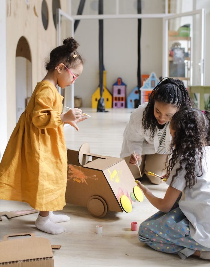 A teen working at day care helping kids paint a cardboard car.